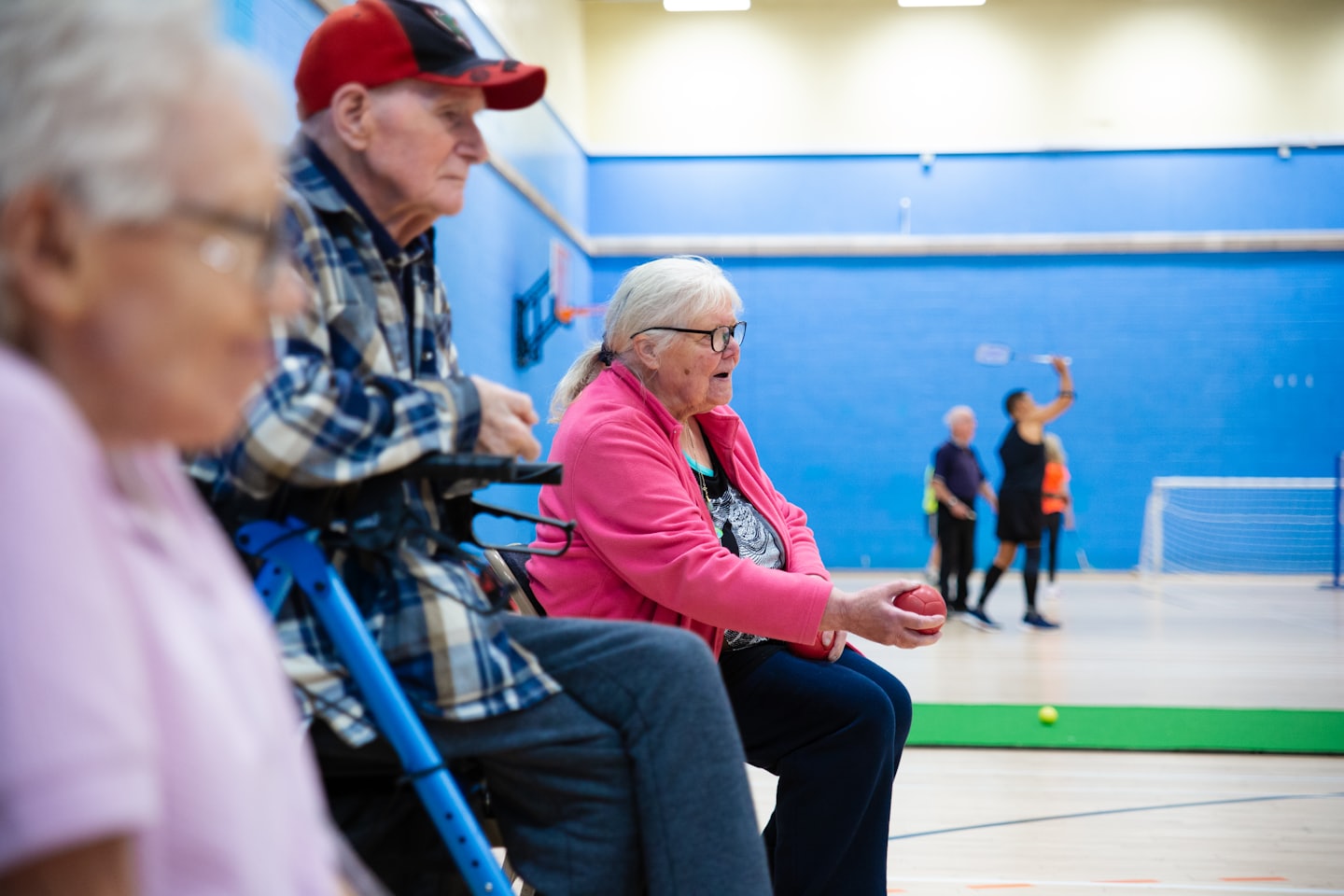 Seniori v telocvični hrajú loptové hry. V pozadí sa hrá bedminton.